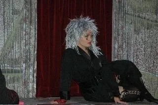 ROCKY HORROR SHOW AT Manoa Valley Theatre. Actress Cassie Favreau-Chung plays Magenta. Woman with spiky silver hair sitting at a table, wearing a black outfit, in front of red curtains.