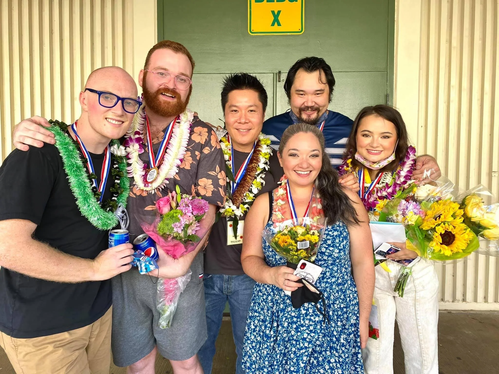 Actors after a show. A group of seven diverse people celebrating together, wearing leis, medals, and holding flowers, with some holding drinks, on a porch or entrance area with a yellow and green sign in the background. 25TH ANNUAL PUTNAM COUNTY