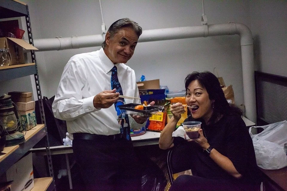 A man in a white dress shirt and tie standing and a woman in black sitting in a room with metal shelves and boxes, enjoying a meal together.