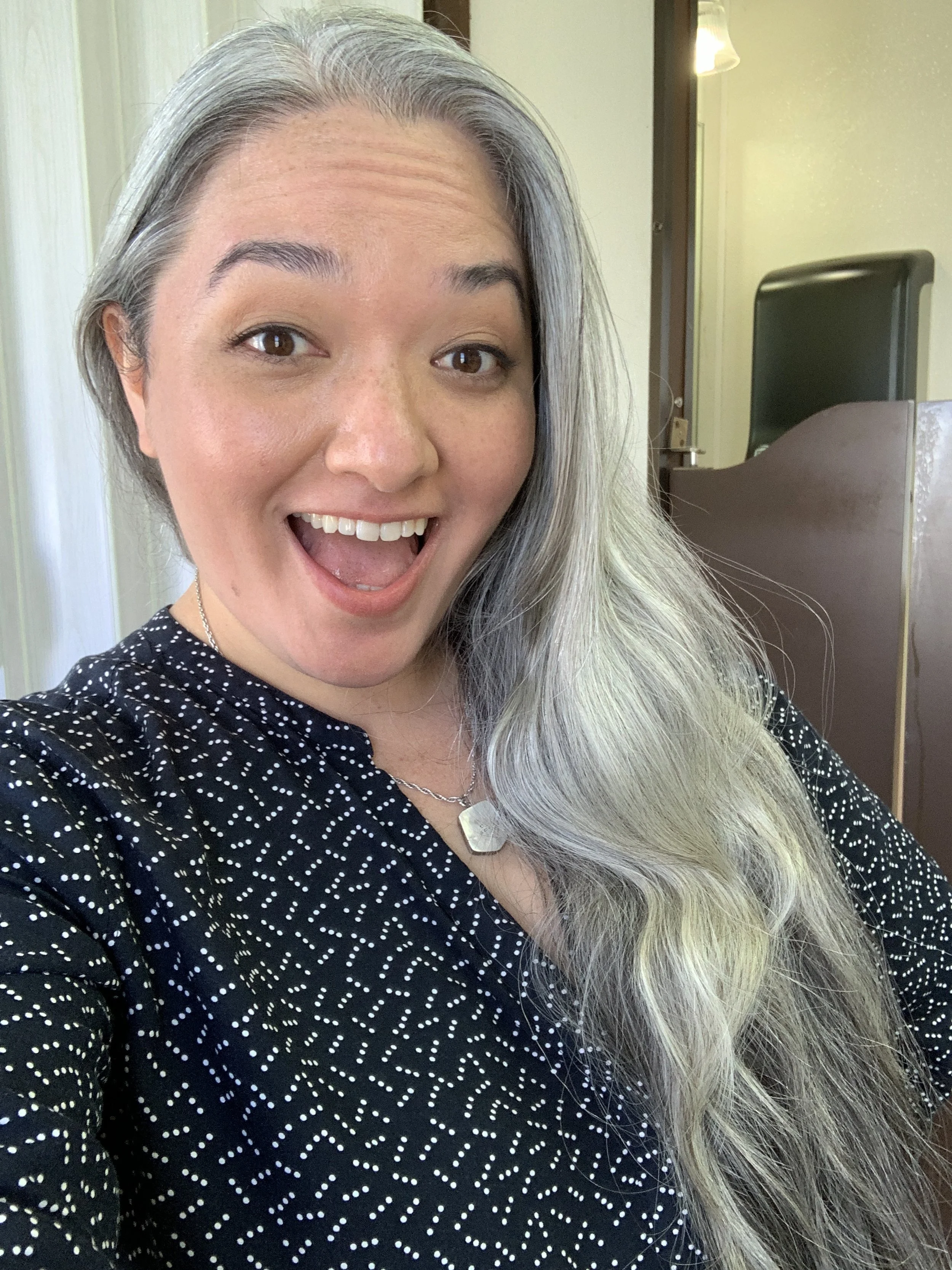 On set filming for MAGNUM P.I. with CBS. A SAG-AFTRA actress taking a selfie, wearing a black and white patterned top and a necklace with a large pendant, with long, silver-blonde hair and a warm indoor setting in the background.