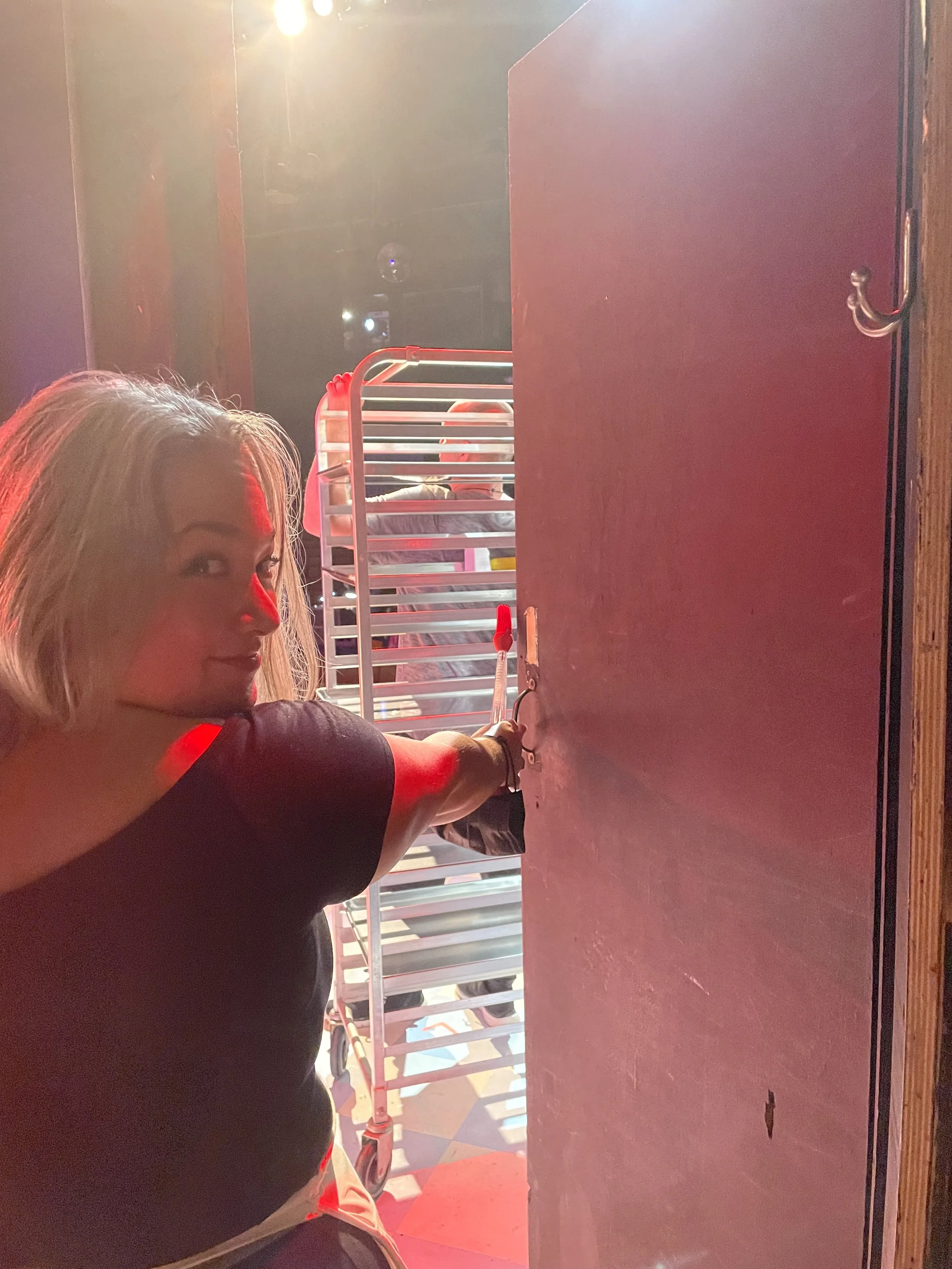 Becky going onstage for "Bad Idea (Reprise)" in Waitress the Broadway musical. Backstage view. A woman with silver hair stands at a door, reaching to open it, with a luggage cart and other people in the background. Red light in theatre.