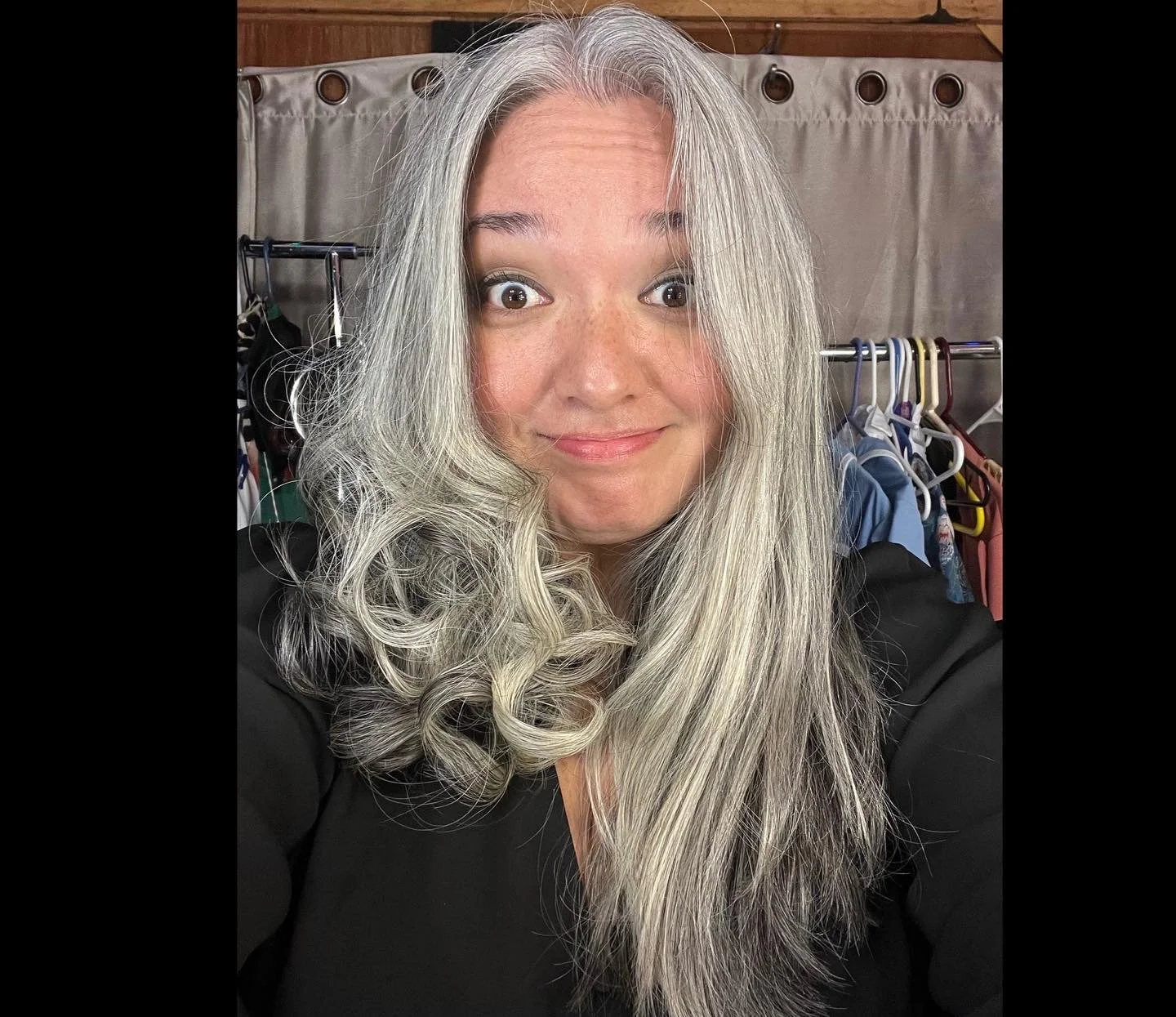 Backstage view in hair and makeup in dressing room. A woman with long, wavy, gray and silver hair smiling at the camera. Behind her, a clothing rack and a beige curtain are visible.