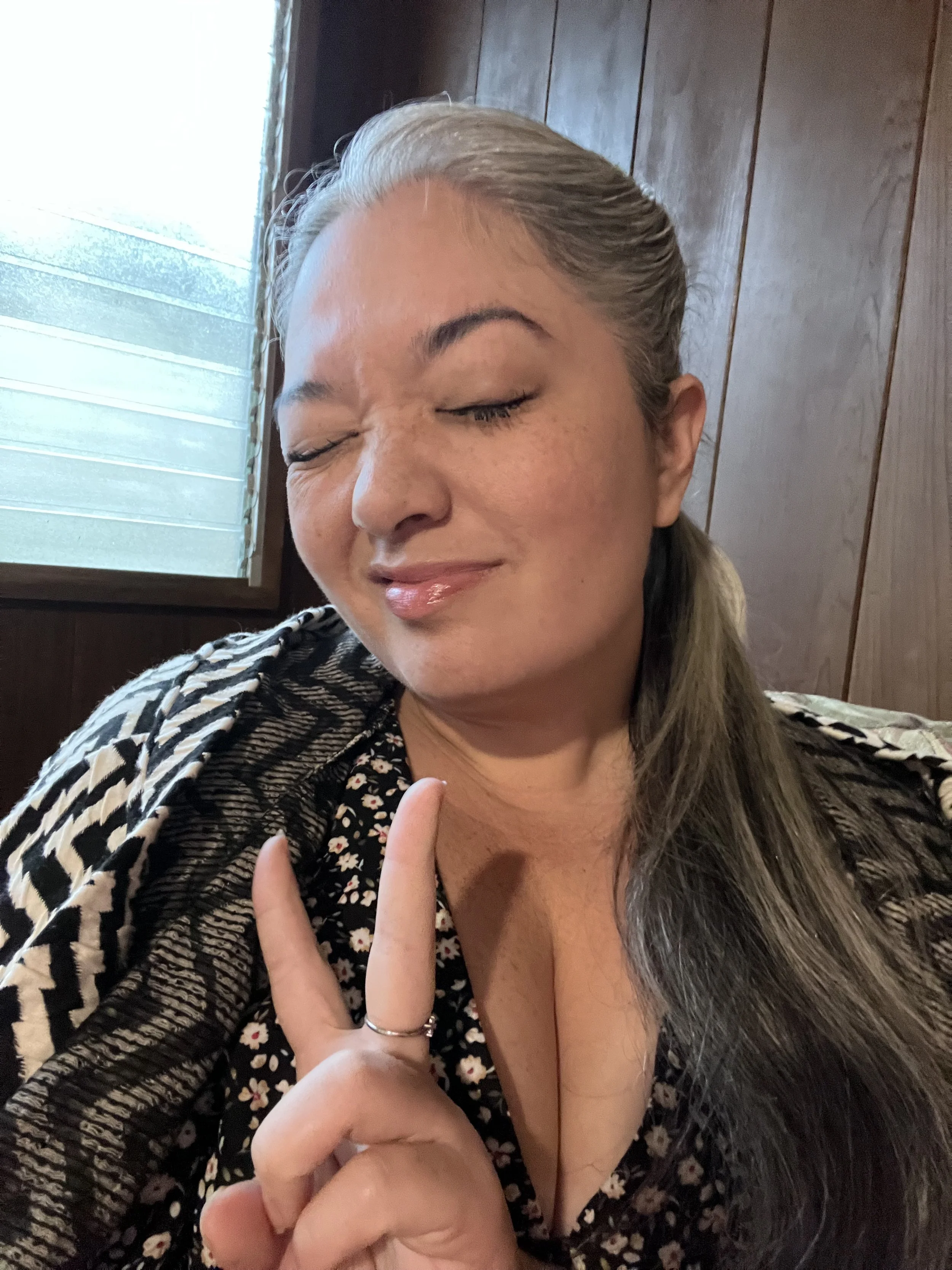 A woman with gray hair and freckles making a peace sign with her hand, eyes closed, smiling softly, sitting indoors near a window with wooden paneling behind her.