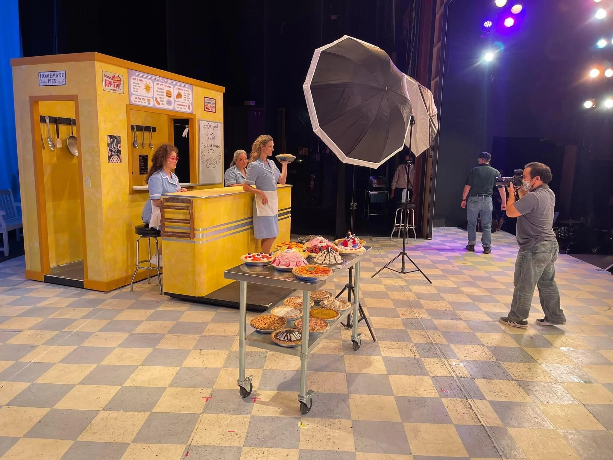 Film set with a yellow diner-themed backdrop, a table with assorted desserts, filming crew with camera, and women in blue uniforms, possibly actors or performers.