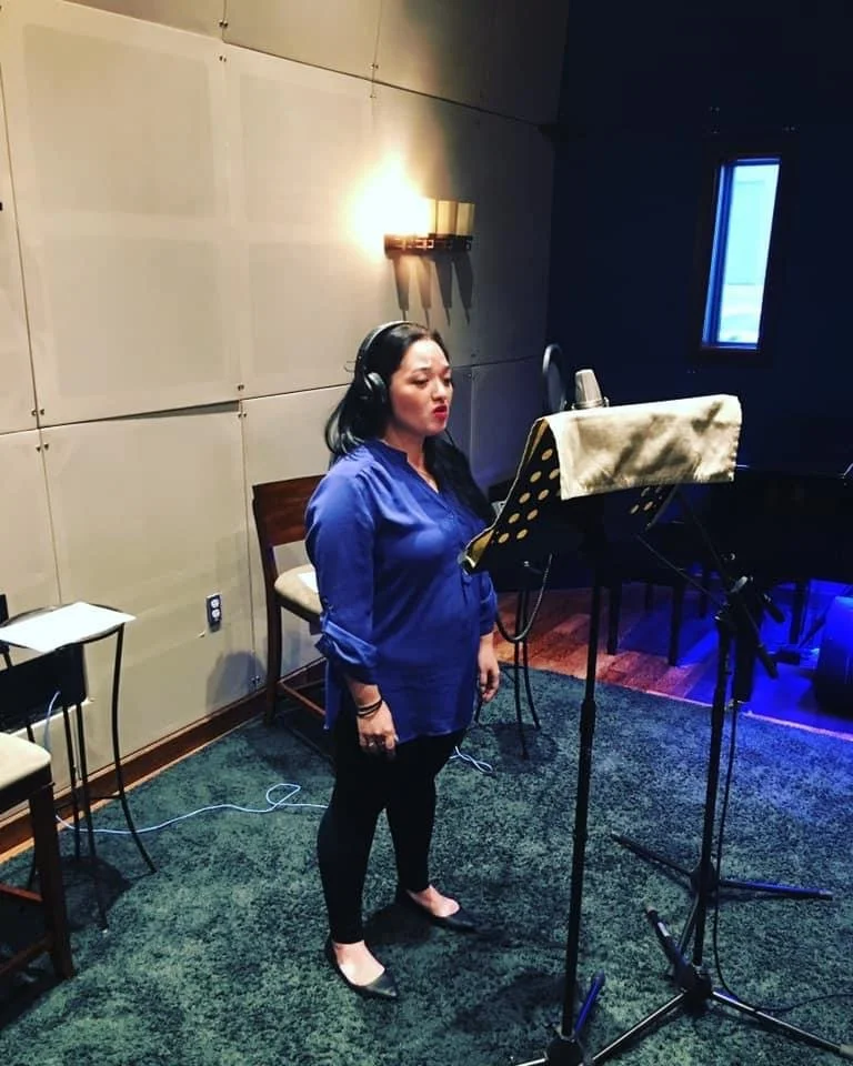 Hawaii SAG-AFTRA voiceover actor. Woman singing in a recording studio, wearing headphones and standing in front of a microphone with a music stand, with acoustic panels on the wall.