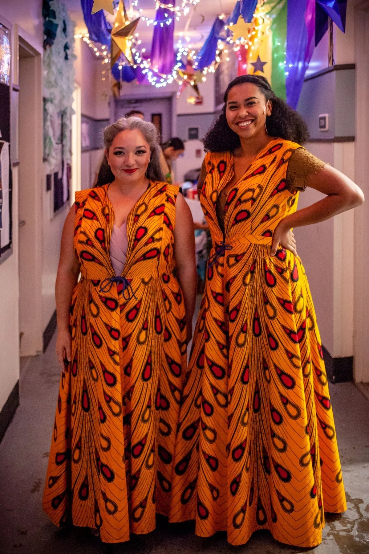 CHILDREN OF EDEN. Two women standing indoors at a celebration, wearing matching vibrant orange dresses with butterfly wing patterns and gold sleeves, decorated with colorful paper stars and streamers hanging from the ceiling, smiling for the camera.