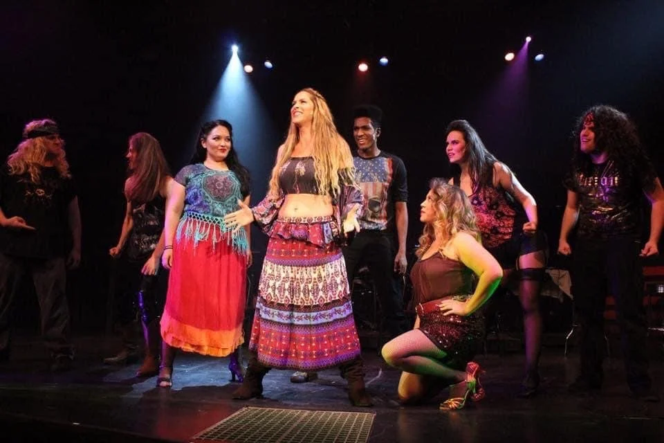 Group of women on stage in colorful, eclectic costumes, performing in a theatrical show with stage lights.