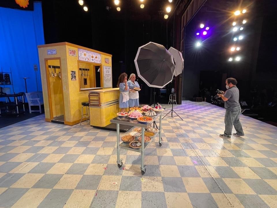 Behind-the-scenes view of a photo shoot set with a small yellow food stand, a table with assorted cakes and baked goods, two women preparing desserts, professional lighting equipment, and a photographer capturing the scene. The floor is checkered, an
