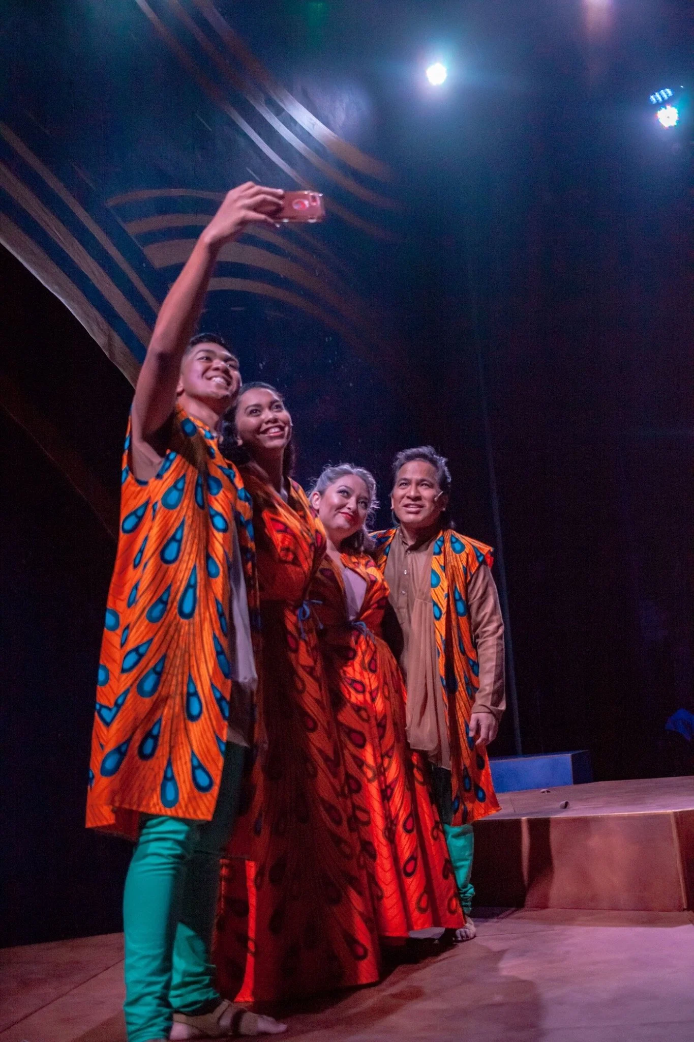 CHILDREN OF EDEN a Broadway musical by I'm A Bright Kid Foundation at Paliku Theatre. Four people wearing traditional colorful clothing taking a group selfie on a stage with a dark background and stage lights.