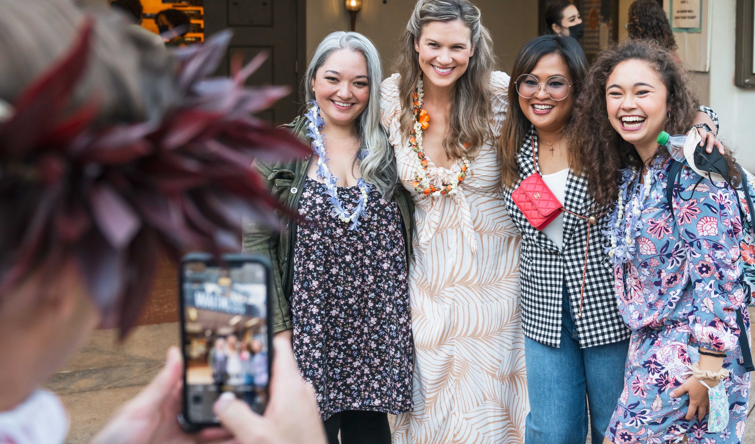 WAITRESS the Broadway musical. Group of five actors standing closely together and smiling at a camera held by a person in front of them. They are outdoors, wearing colorful and patterned clothing, with some wearing floral leis and accessories.