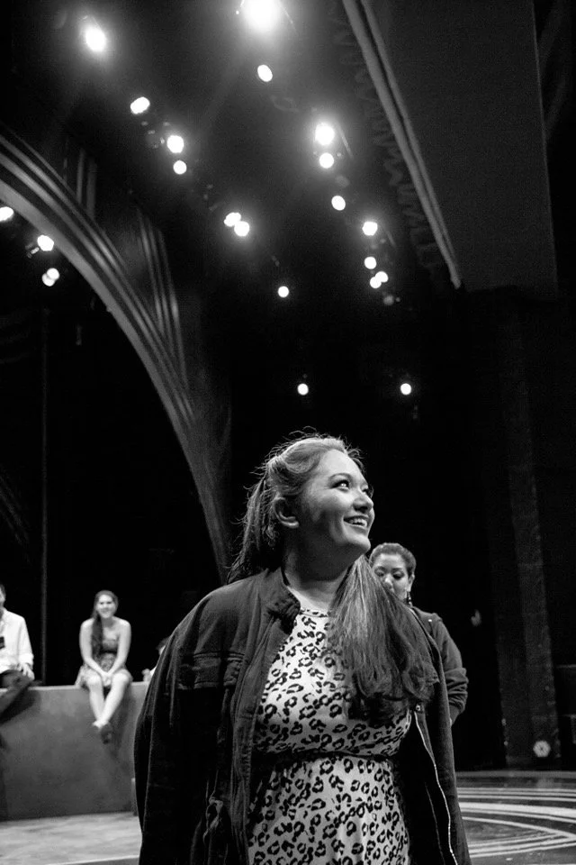 CHILDREN OF EDEN a Broadway musical by I'm A Bright Kid Foundation at Paliku Theatre. A woman smiling and looking to the side on a stage or theater, with stage lighting above and other people sitting or standing in the background.