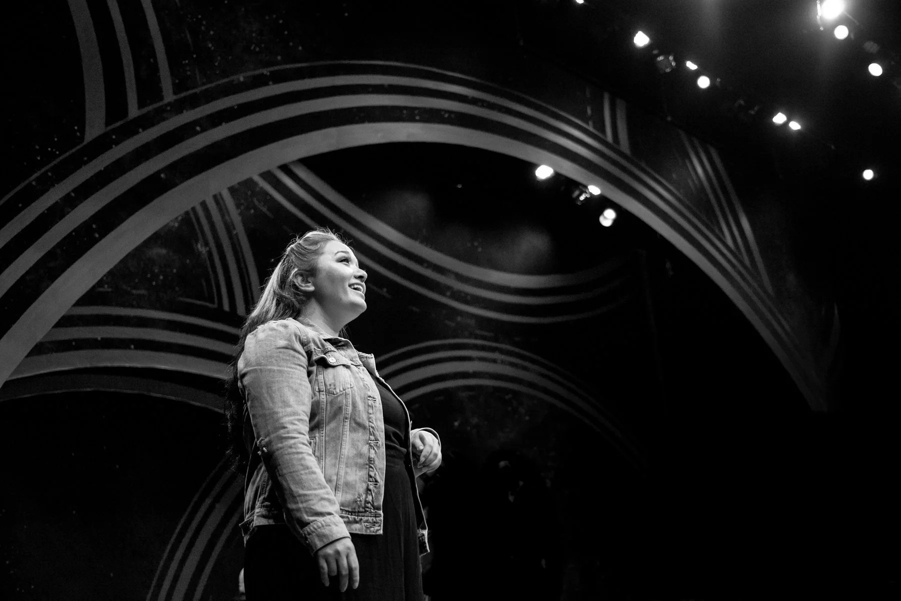 CHILDREN OF EDEN a Broadway musical by I'm A Bright Kid Foundation at Paliku Theatre. A woman smiling and looking up on a stage with curved graphic designs in the background, in black and white.