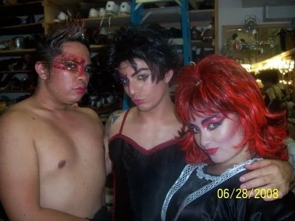 ROCKY HORROR SHOW AT Manoa Valley Theatre. Actress Cassie Favreau-Chung plays Magenta. Three drag performers in a backstage area with shelves of costumes and props, dressed in elaborate makeup and costumes, standing close together.