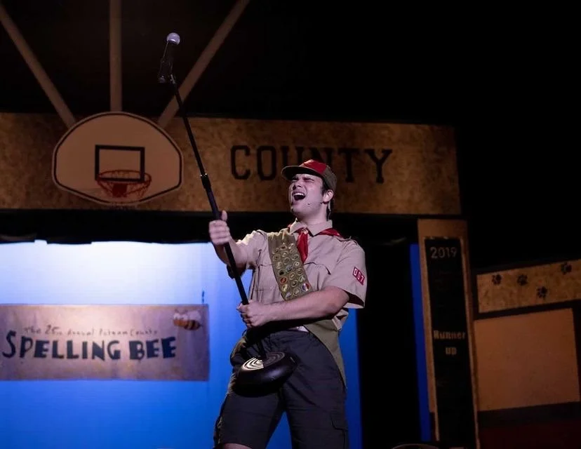 THE 25TH ANNUAL PUTNAM COUNTY SPELLING BEE a comedy musical at Manoa Valley Theatre in Honolulu, Hawaii. A young man dressed as a scout holding a golf club, wearing a tan uniform with a red neckerchief, smiling on stage at the spelling bee event.