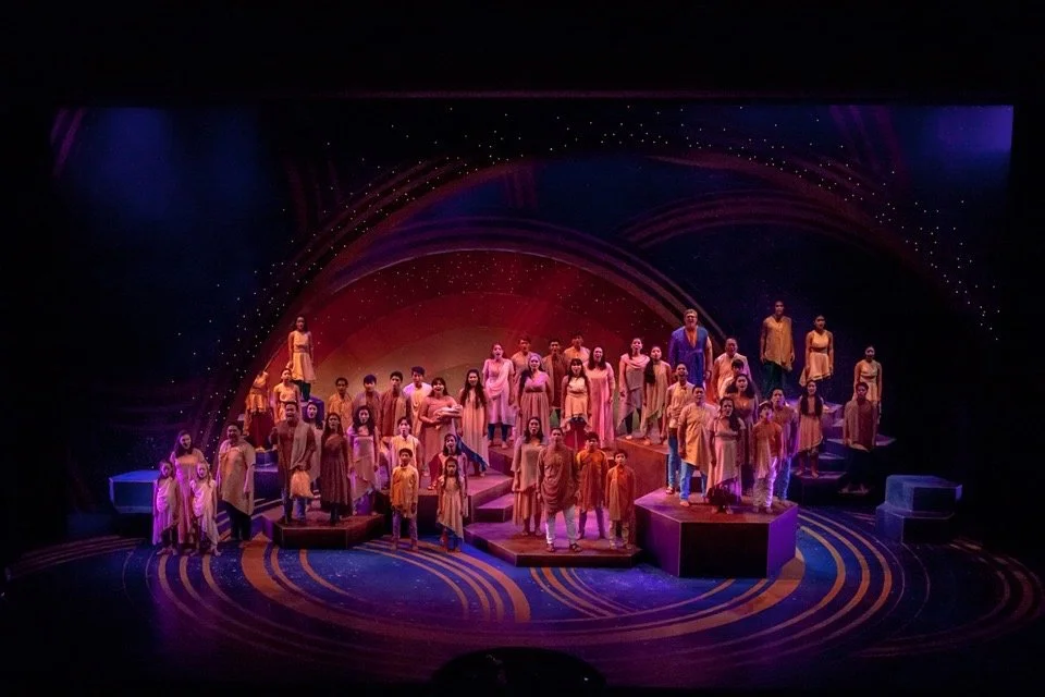 CHILDREN OF EDEN a Broadway musical by I'm A Bright Kid Foundation at Paliku Theatre. A large group of performers dressed in yellow and orange costumes on a colorful stage during a theatrical production.