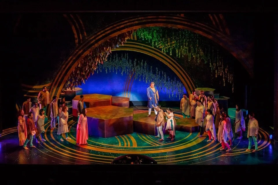 A theatrical stage with actors performing in colorful costumes, with a vibrant background and stage design including rings and hanging decorations.