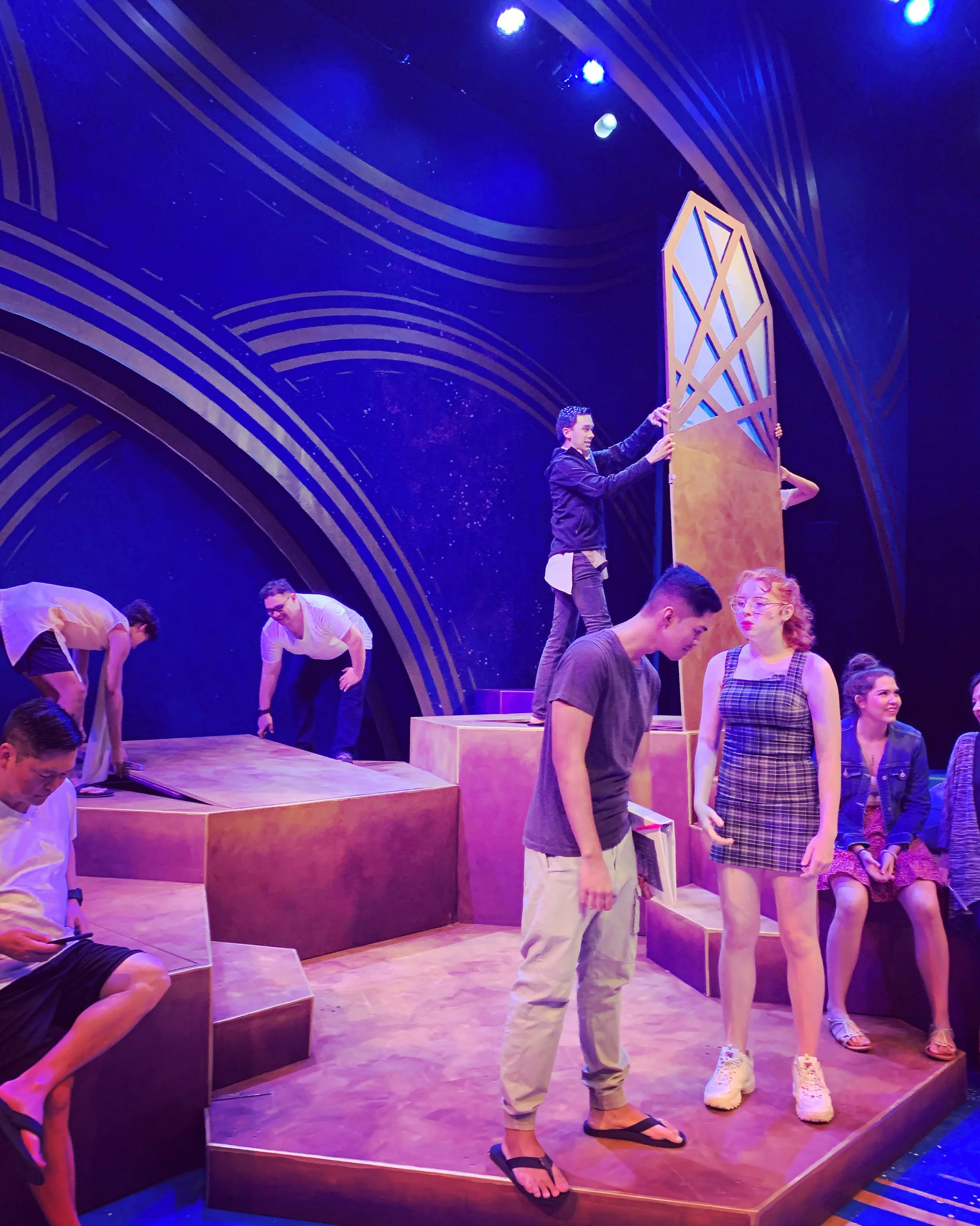 CHILDREN OF EDEN. The stage of a theater with multiple performers practicing or performing. Some are adjusting or setting up scenery, while others stand and wait. The background features a dark blue backdrop with gold curved lines and lighting.
