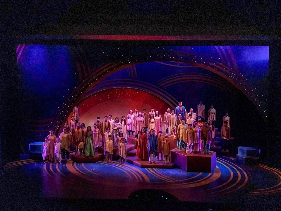 CHILDREN OF EDEN a Broadway musical by I'm A Bright Kid Foundation at Paliku Theatre. Stage performance featuring a large cast of singers dressed in colorful costumes, standing on tiered platforms with a vibrant, abstract backdrop and stage lighting.