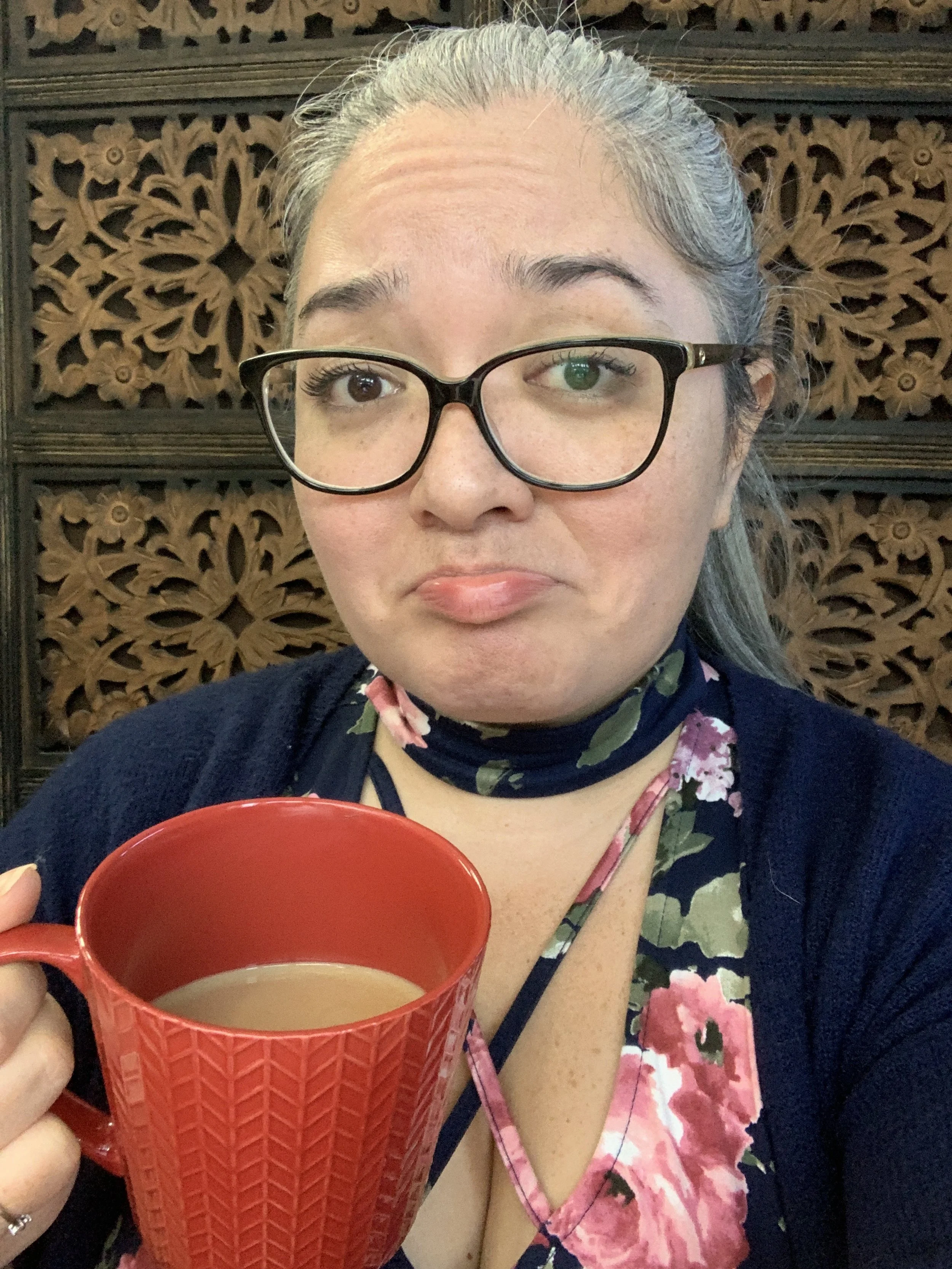 A woman with glasses and gray hair is holding a red mug and looking at the camera, with a wooden decorative background behind her.