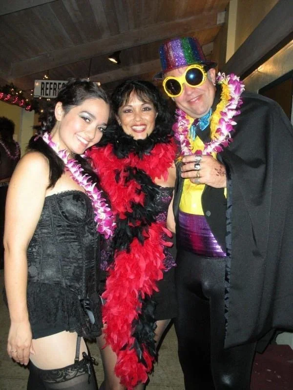 ROCKY HORROR SHOW AT Manoa Valley Theatre. Actress Cassie Favreau-Chung plays Magenta. Three people dressed in colorful, festive costumes at a party, smiling and wearing accessories such as leis, a feather boa, sunglasses, and a colorful hat.