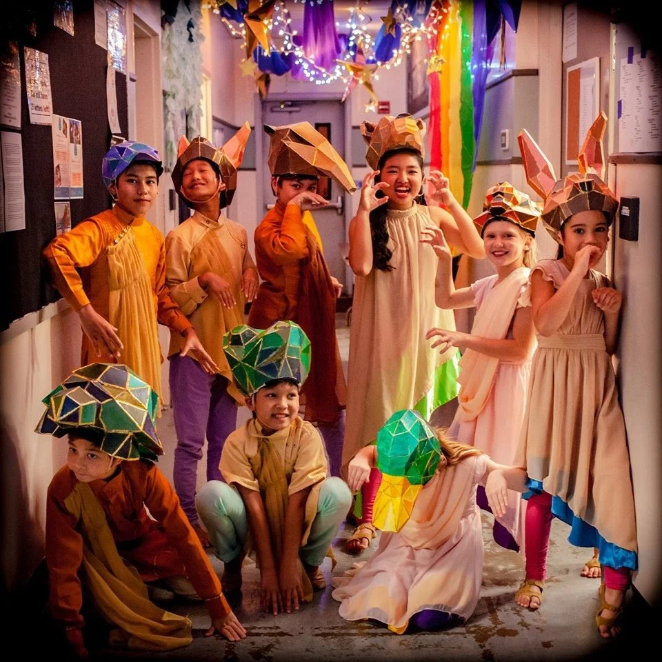 Children dressed in animal costumes, some wearing paper hats shaped like foxes and other animals, in a decorated hallway with colorful streamers and lights.