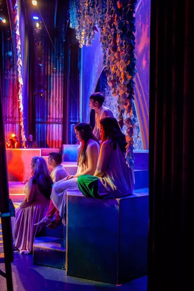 CHILDREN OF EDEN a Broadway musical by I'm A Bright Kid Foundation at Paliku Theatre. Group of young women and a young man sitting and standing on a stage or platform, watching a performance in a colorful, decorated theater with curtains and foliage.