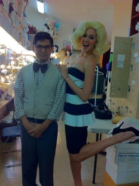 LITTLE SHOP OF HORRORS a comedic musical at Manoa Valley. A woman dressed like a glamorous celebrity with a big blonde wig, posing and smiling next to a man in a dress shirt, bow tie, and vest at what appears to be a thrift store or costume shop.