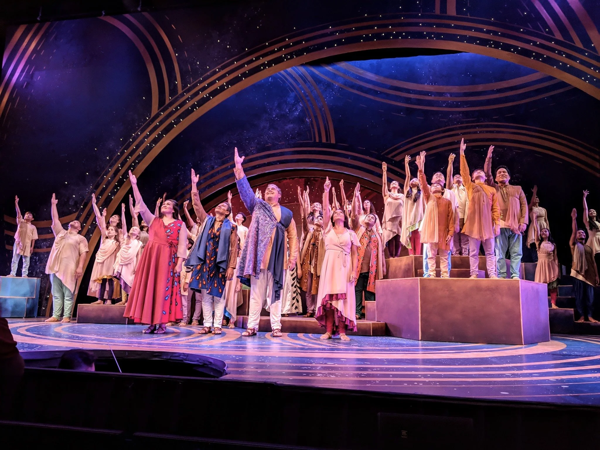 CHILDREN OF EDEN a Broadway musical by I'm A Bright Kid Foundation at Paliku Theatre. A large group of performers on a stage with a cosmic-themed backdrop, standing on tiered platforms, raising one arm upward in a synchronized gesture.
