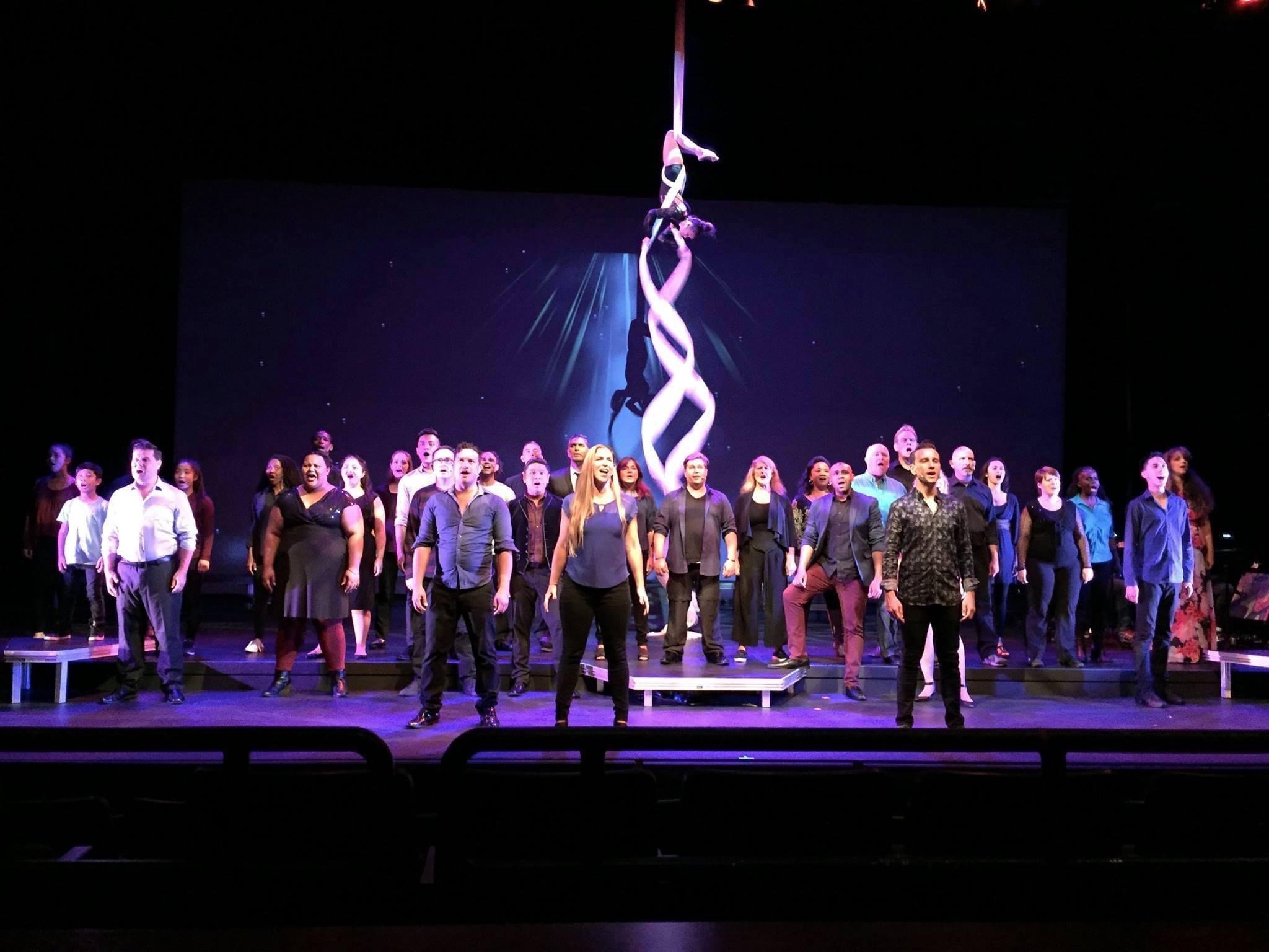 RISE UP: A BENEFIT CONCERT BY MELINA LILLIOS at Paliku Theatre in Kaneohe, Hawaii. Large group of diverse performers singing on stage with projected background of aerial acrobats twirling in the air.