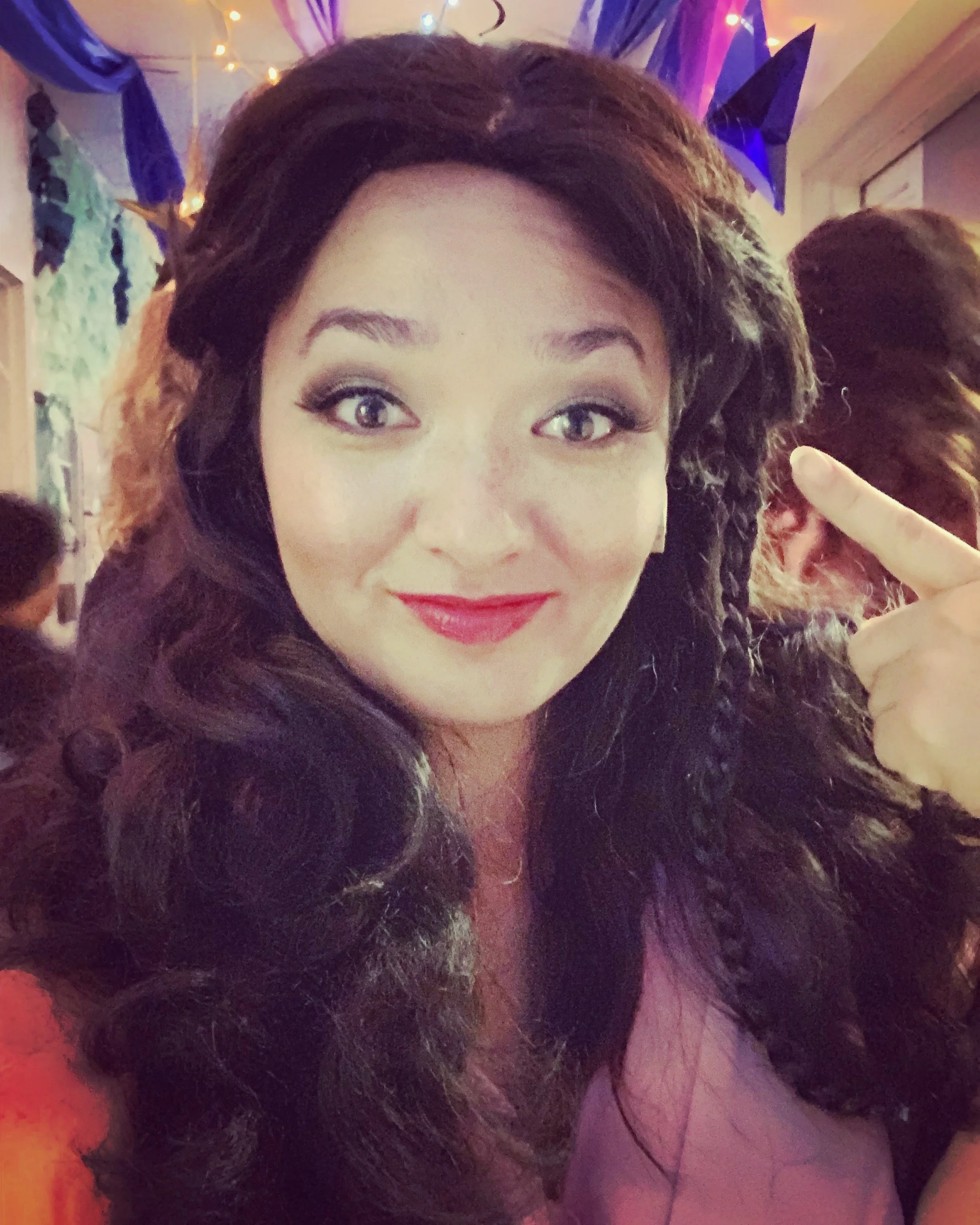 CHILDREN OF EDEN a Broadway musical by I'm A Bright Kid Foundation at Paliku. A woman with long curly hair, wearing makeup with red lipstick, pointing to her head wig, in a festive setting with colorful decorations and other people in the background.