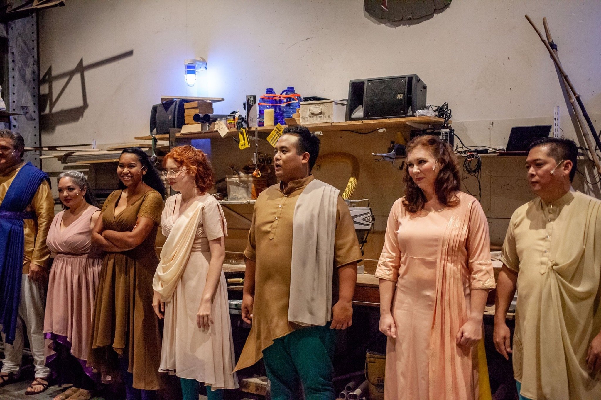 CHILDREN OF EDEN a Broadway musical by I'm A Bright Kid Foundation at Paliku Theatre. Group of seven actors in traditional Southeast Asian costumes standing in a line, behind them is a workshop with shelves, tools, and equipment.