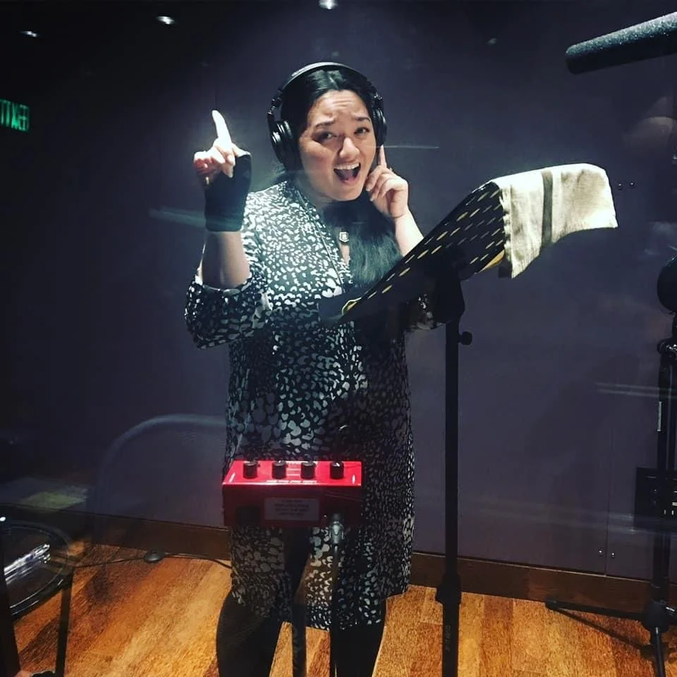 A woman singing in a recording studio, wearing headphones and a patterned dress, standing in front of a microphone with a music stand and sheet, raising her right index finger and smiling. Hawaii voiceover actor at Blue Planet Sound in Honolulu.