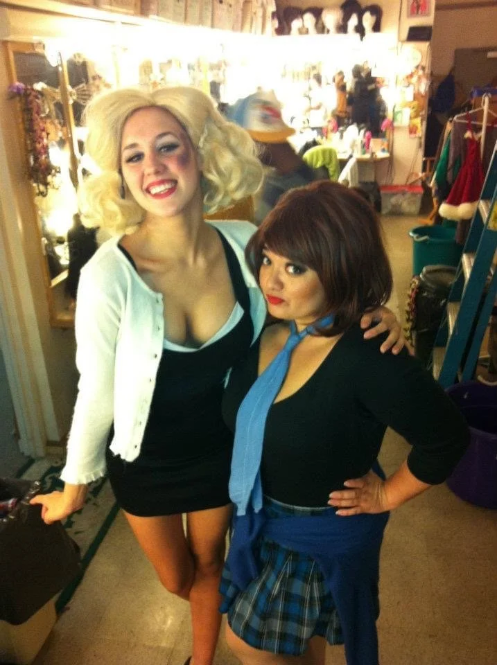 LITTLE SHOP OF HORRORS a comedic musical at Manoa Valley Theatre in Honolulu, Hawaii. Two women in costumes standing inside a store, one with blonde hair and a white cardigan, the other with short brown hair in a black top and blue plaid skirt.