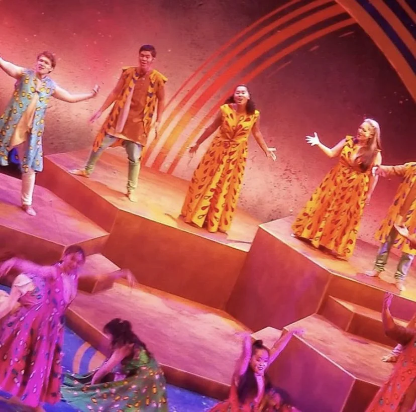 CHILDREN OF EDEN a Broadway musical by I'm A Bright Kid Foundation at Paliku Theatre.Group of performers on a stage dressed in colorful costumes, some standing on raised platforms, with a vibrant background featuring curved red lines.