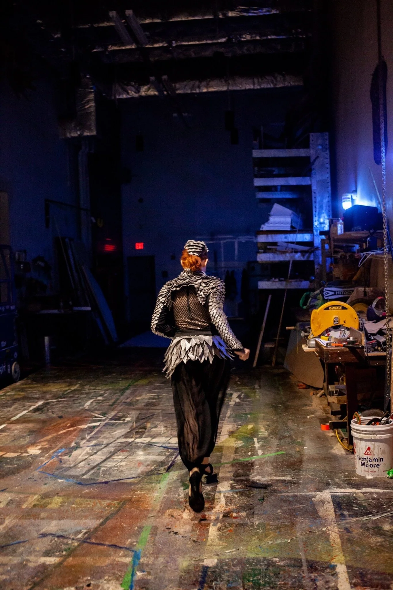 A woman dressed in artistic, elaborate black and white costume, walking away in a dimly lit, cluttered workshop or studio with tools and materials around.
