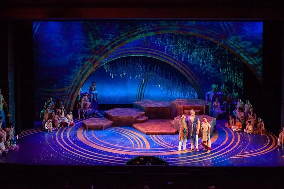 The image depicts a stage performance with actors, some standing in the center and others seated on multi-level platforms and the floor. The backdrop features a digital or painted design with blue and gold colors and abstract patterns.