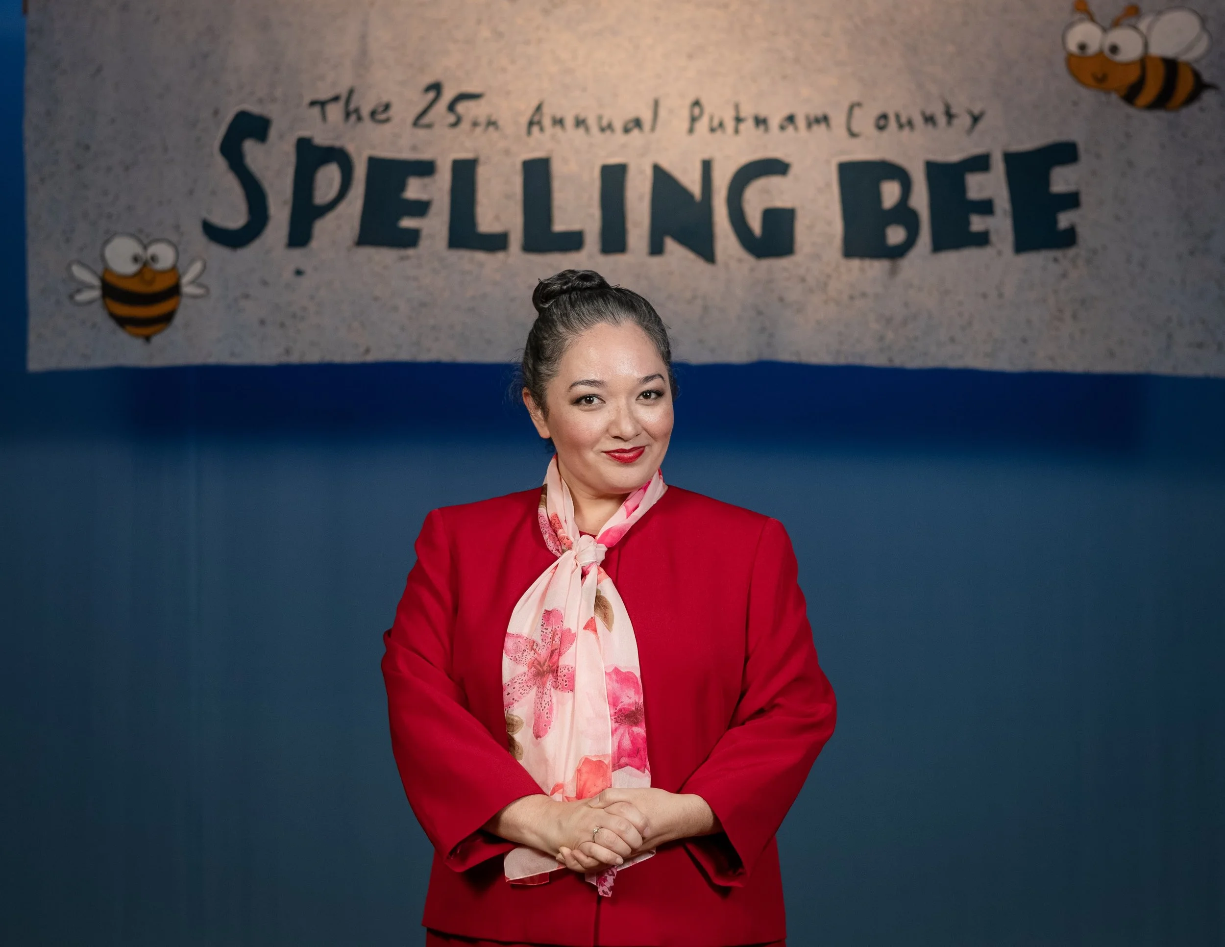 A woman in a red blazer and pink floral scarf standing in front of a banner that reads 'The 25th Annual Putnam County Spelling Bee' with cartoon bees on it.