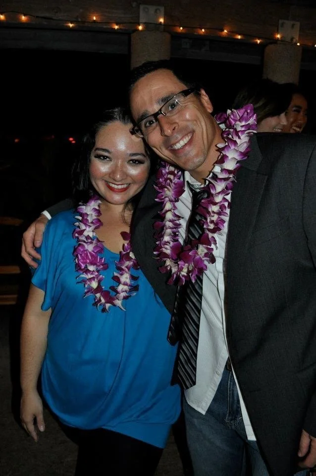 LITTLE SHOP OF HORRORS a comedic musical at Manoa Valley Theatre in Honolulu, Hawaii. Two people smiling and wearing purple and white leis at a celebration or party, with others in the background.