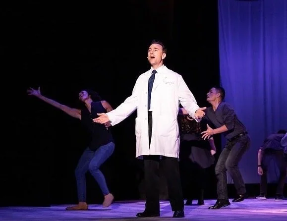 WAITRESS the Broadway musical by Sara Bareilles at Diamond Head Theatre. A theatrical performance featuring a Zack Linnert in a white lab coat, who appears to be acting or singing on stage, with backup dancers or actors in the background.
