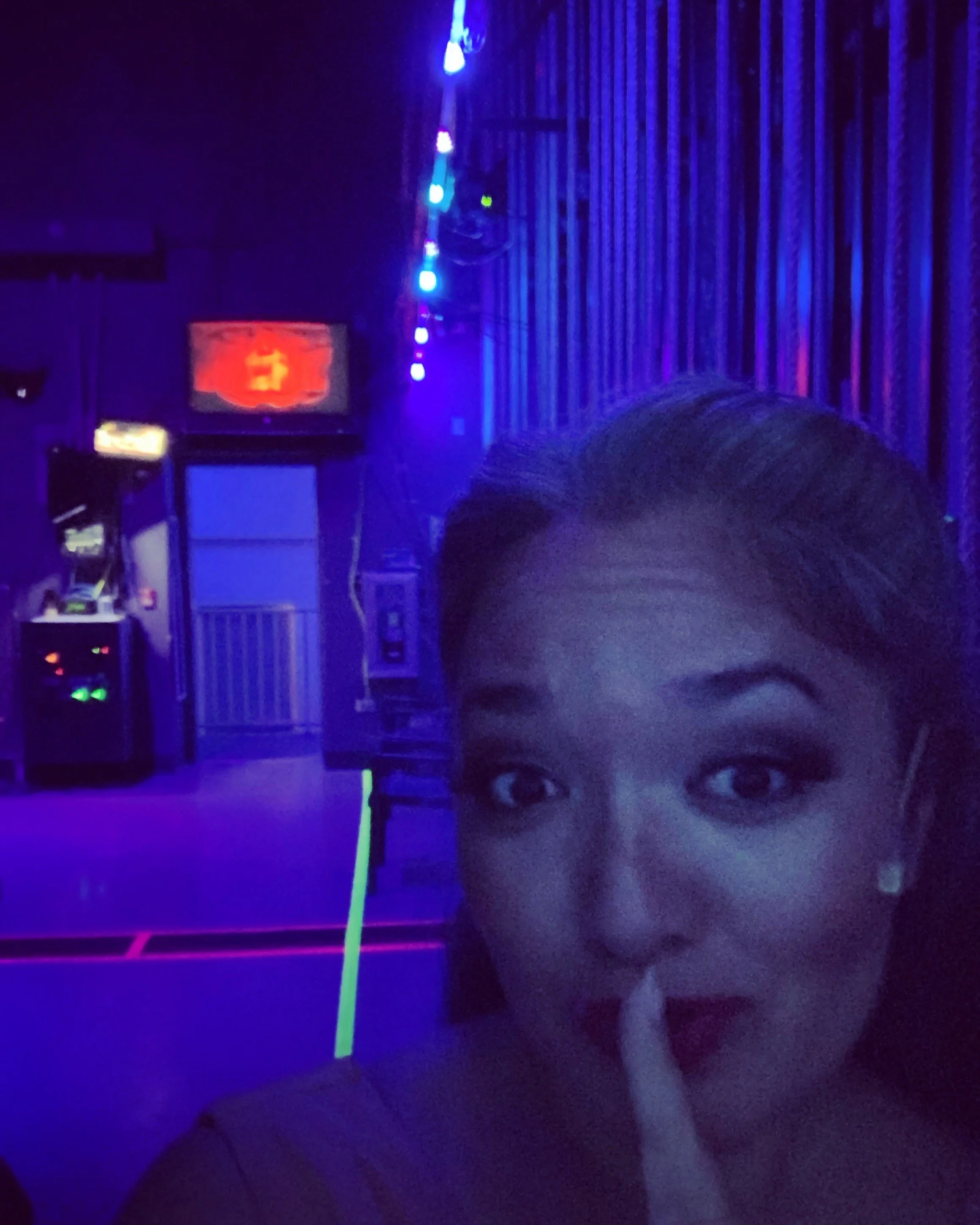 CHILDREN OF EDEN a Broadway musical by I'm A Bright Kid Foundation at Paliku Theatre. A woman taking a selfie in a dark, neon-lit arcade or entertainment venue, with blue and purple lighting, a large screen in the background, and arcade machines.