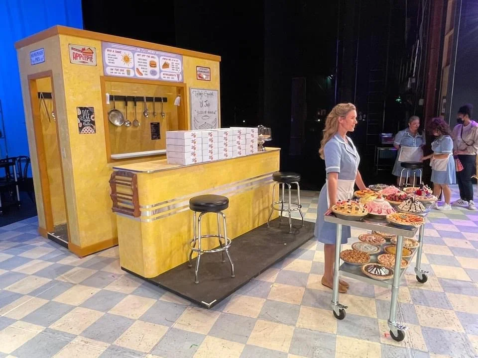 A woman standing behind a table full of assorted cookies and baked goods at a bakery or cafe set up. In the background, there are other people, some in uniforms, and a yellow wooden counter with boxes on it.