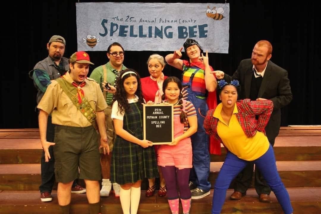 Group of children and adults on stage at the Putnam County Spelling Bee event, with a banner in the background reading 'The 25th Annual Putnam County Spelling Bee' and a girl in the front holding a sign that says '25th Annual Putnam County Spelling B