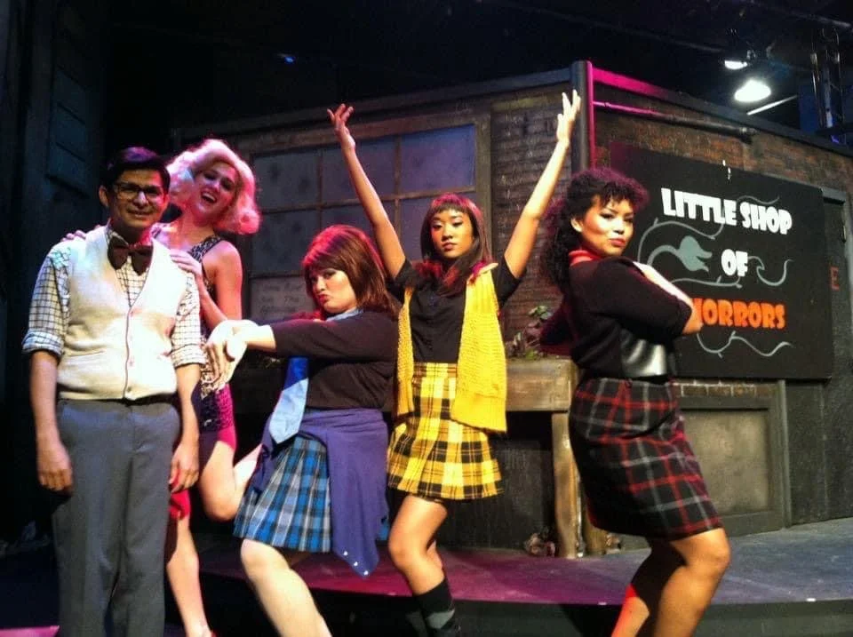 The image shows five people on stage, two men and three women, in front of a sign that says 'Little Shop of Horrors.' The performers are dressed in colorful, 1950s-inspired costumes and are striking playful poses, with one woman raising her arms and 
