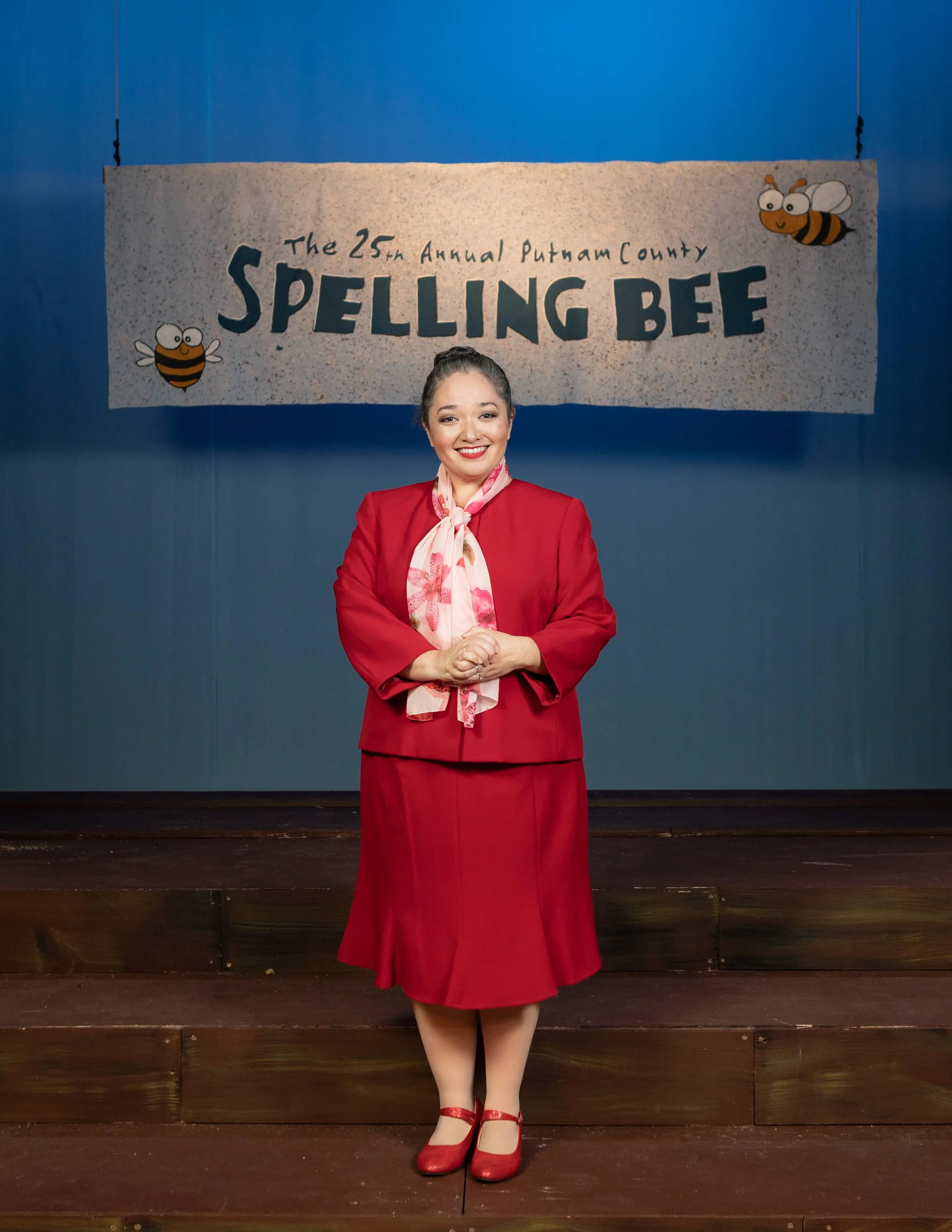 THE 25TH ANNUAL PUTNAM COUNTY SPELLING BEE a comedy musical at Manoa Valley. A woman in a red skirt suit with a pink floral scarf stands on stage in front of a sign that reads 'The 25th Annual Putnam County Spelling Bee' with animated bee characters.