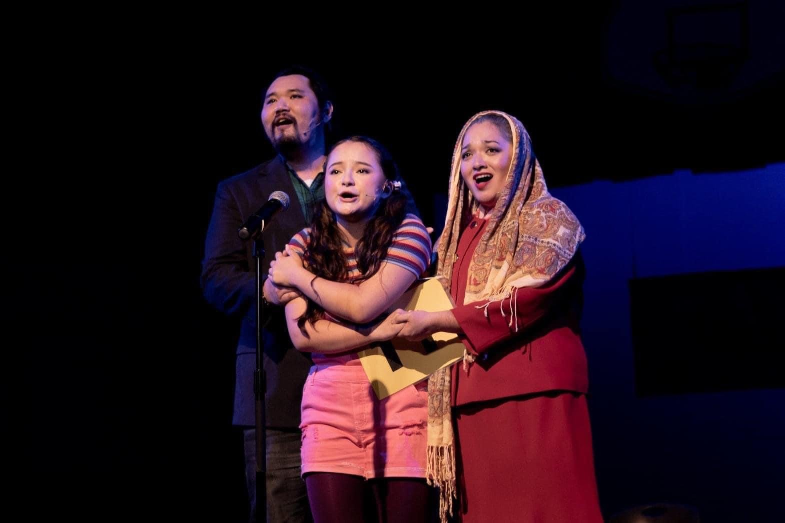 THE 25TH ANNUAL PUTNAM COUNTY SPELLING BEE a comedy musical at Manoa Valley Theatre in Honolulu, Hawaii. Three people on stage singing and acting, with one holding a yellow box, under stage lighting, against a dark background. "The I Love You Song"