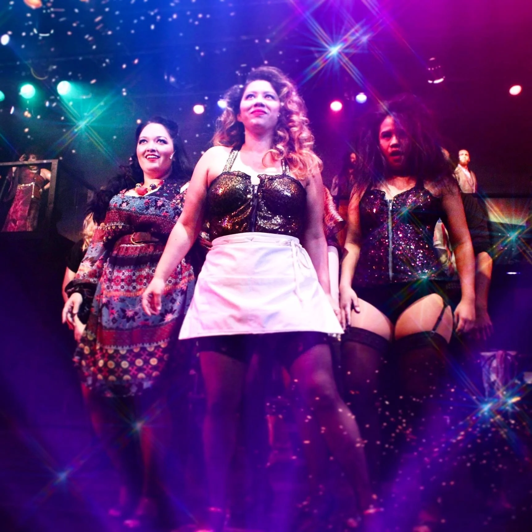 ROCK OF AGES a rock musical at Manoa Valley Theatre. Three women walking confidently on stage under colorful lights, with one of them dressed as a waitress with a pink apron and black top, others wearing revealing outfits, energetic atmosphere.