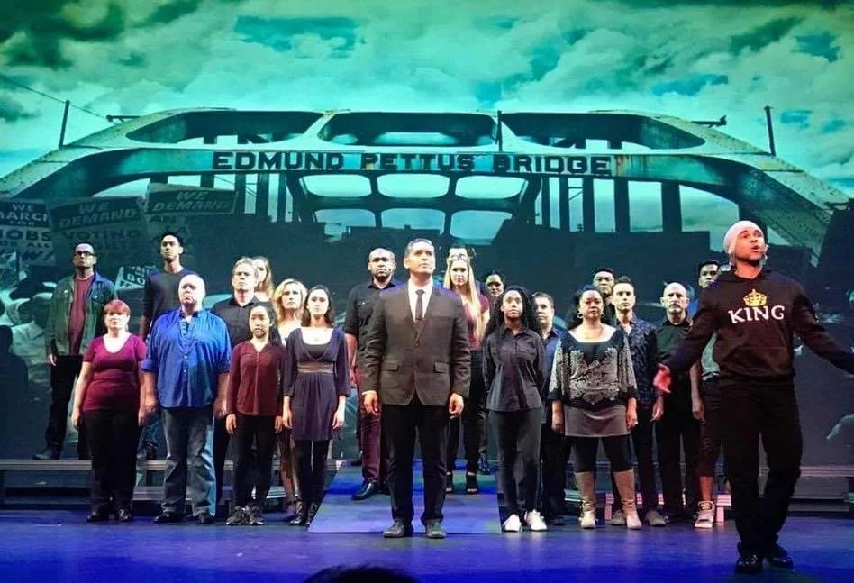 RISE UP: A BENEFIT CONCERT BY MELINA LILLIOS at Paliku Theatre in Kaneohe, Hawaii. A group of people performing on a stage with a backdrop of the Edmund Pettus Bridge in Selma, Alabama, during a theatrical production or reenactment.
