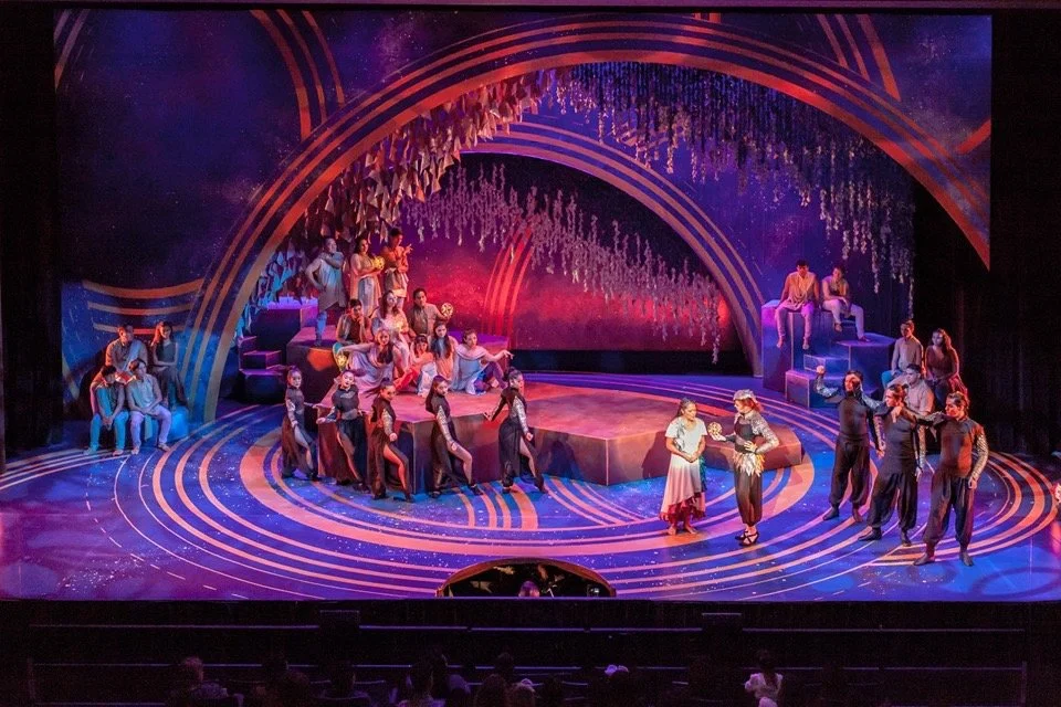 On stage, a theatrical performance with numerous actors and actresses. The set features colorful, curved patterns and hanging decorations with performers seated or standing, suggesting a dramatic or musical scene.