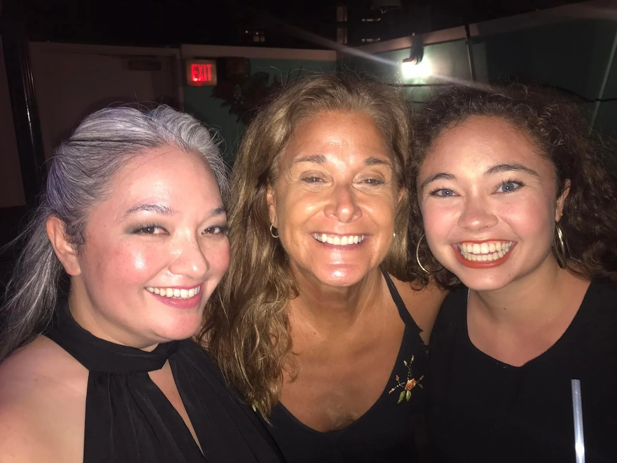 Hawaii performers, musicians and actresses. Three women smiling and posing together at a social event in a dimly lit indoor setting.