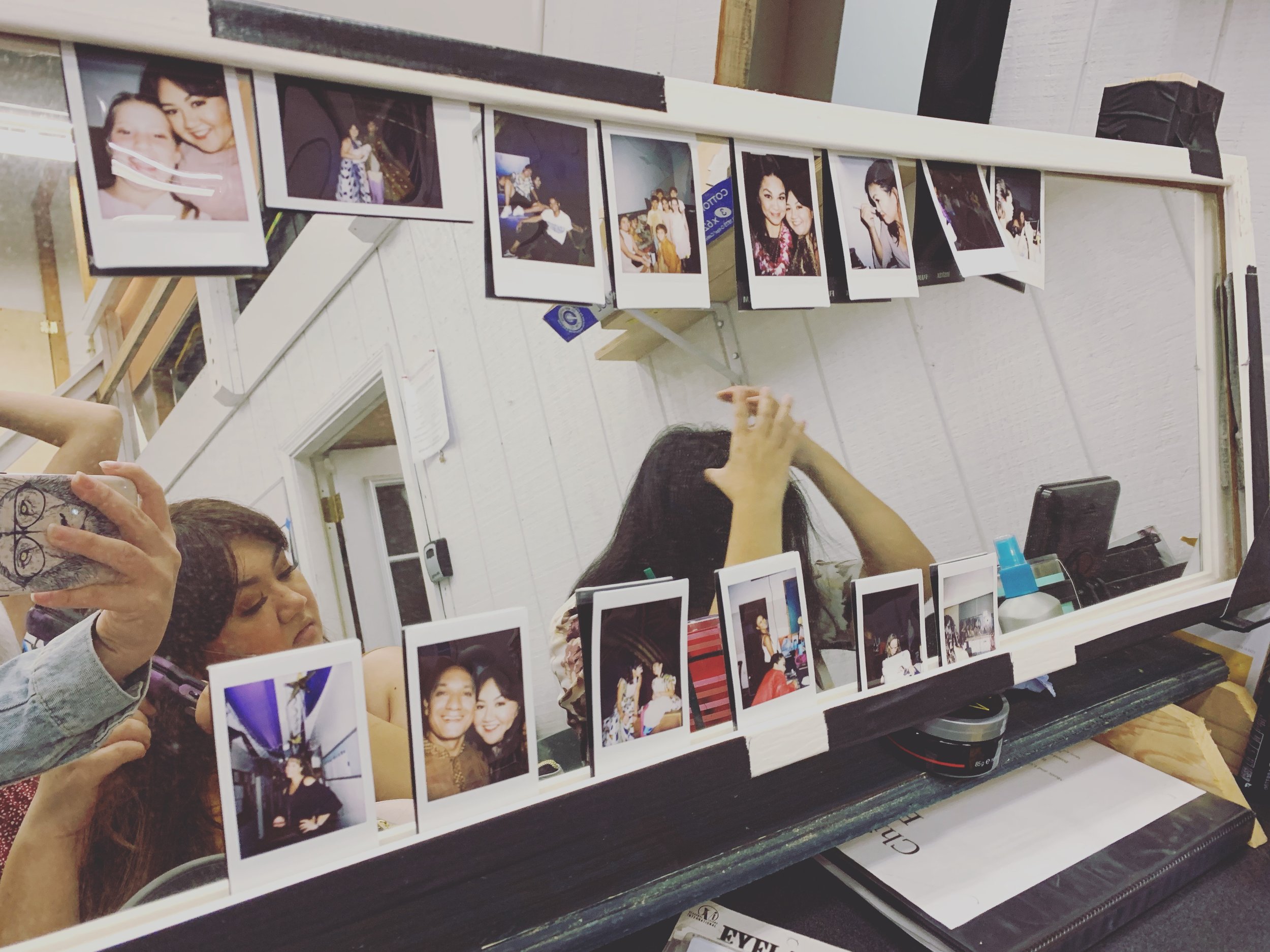 CHILDREN OF EDEN a Broadway musical by I'm A Bright Kid Foundation at Paliku Theatre. Reflection of two women taking a selfie in a mirror surrounded by printed photographs hanging on a string, with a white wall in the background.